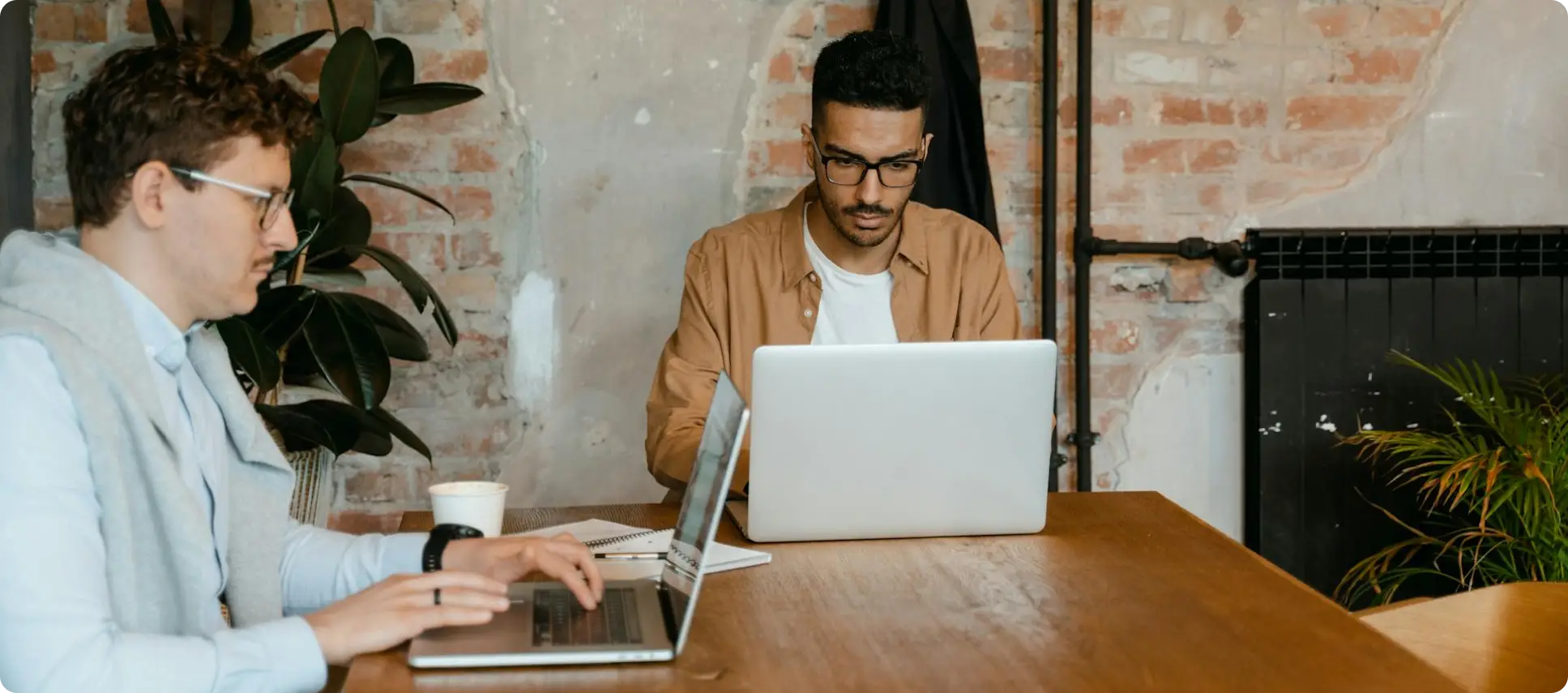 Two male developers at Changeable working on laptops in a focused New Plymouth office environment. Featuring exposed brick and natural wood, this image represents the technical execution and collaborative deep work of AI implementation in Taranaki, NZ.